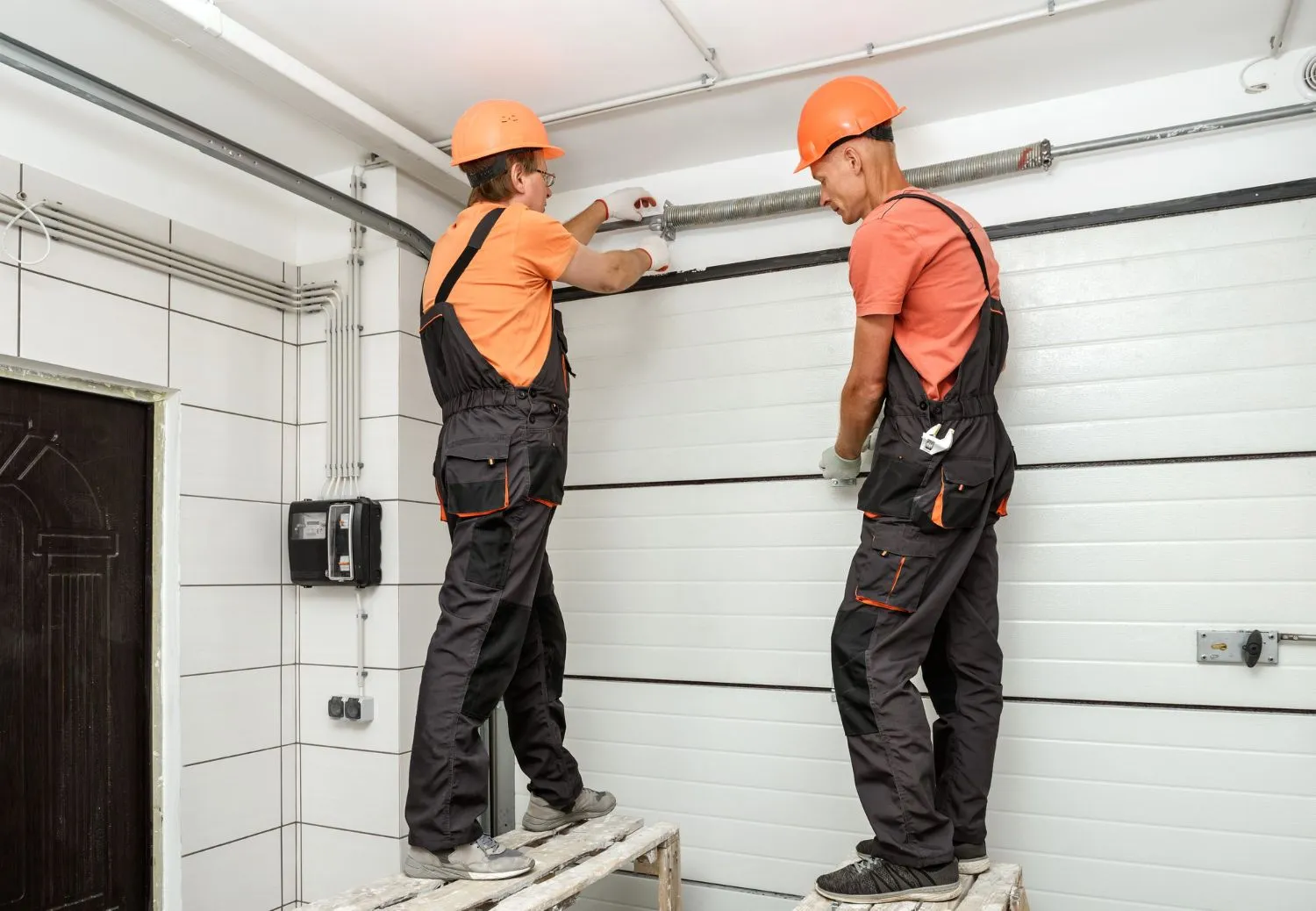 Two technicians on trestles fitting the torsion spring above a white sectional door